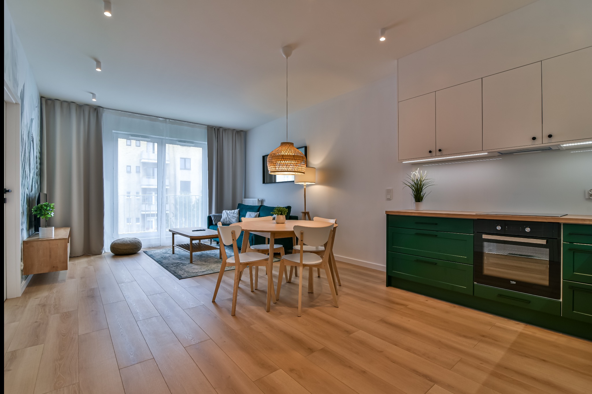 Nowoczesne wnętrze: salon z aneksem kuchennym. Drewniana podłoga, stół z krzesłami, zielona zabudowa kuchenna i wygodna sofa. Jasne, przestronne mieszkanie.