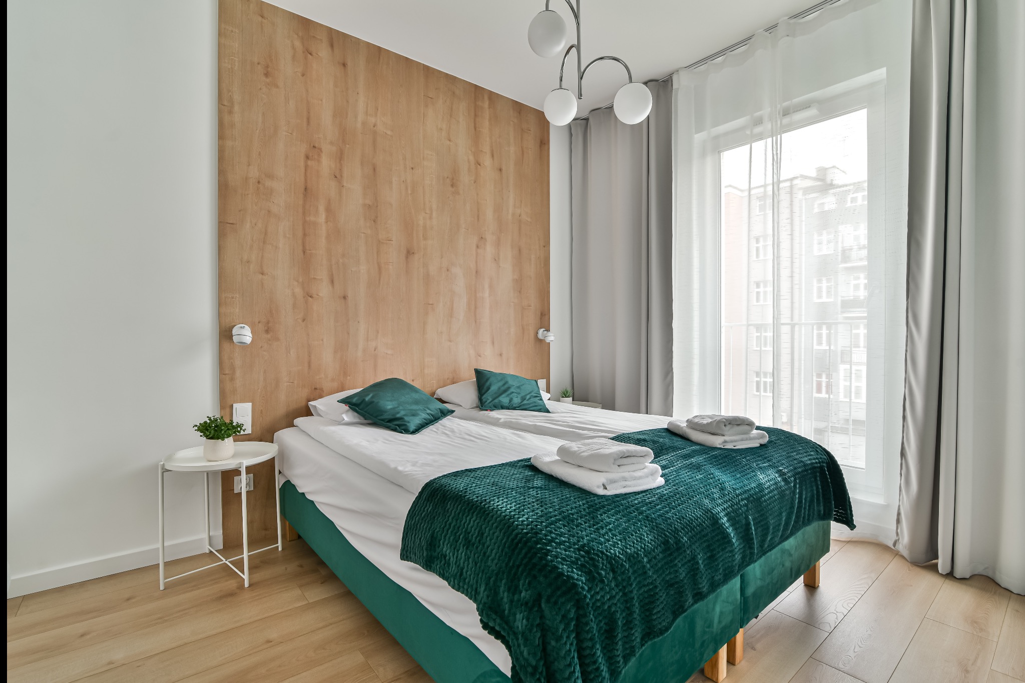 Jasna sypialnia z drewnianą ścianą, dwoma łóżkami z zielonymi akcentami i białymi ręcznikami. Widok na budynek przez okno z firankami.