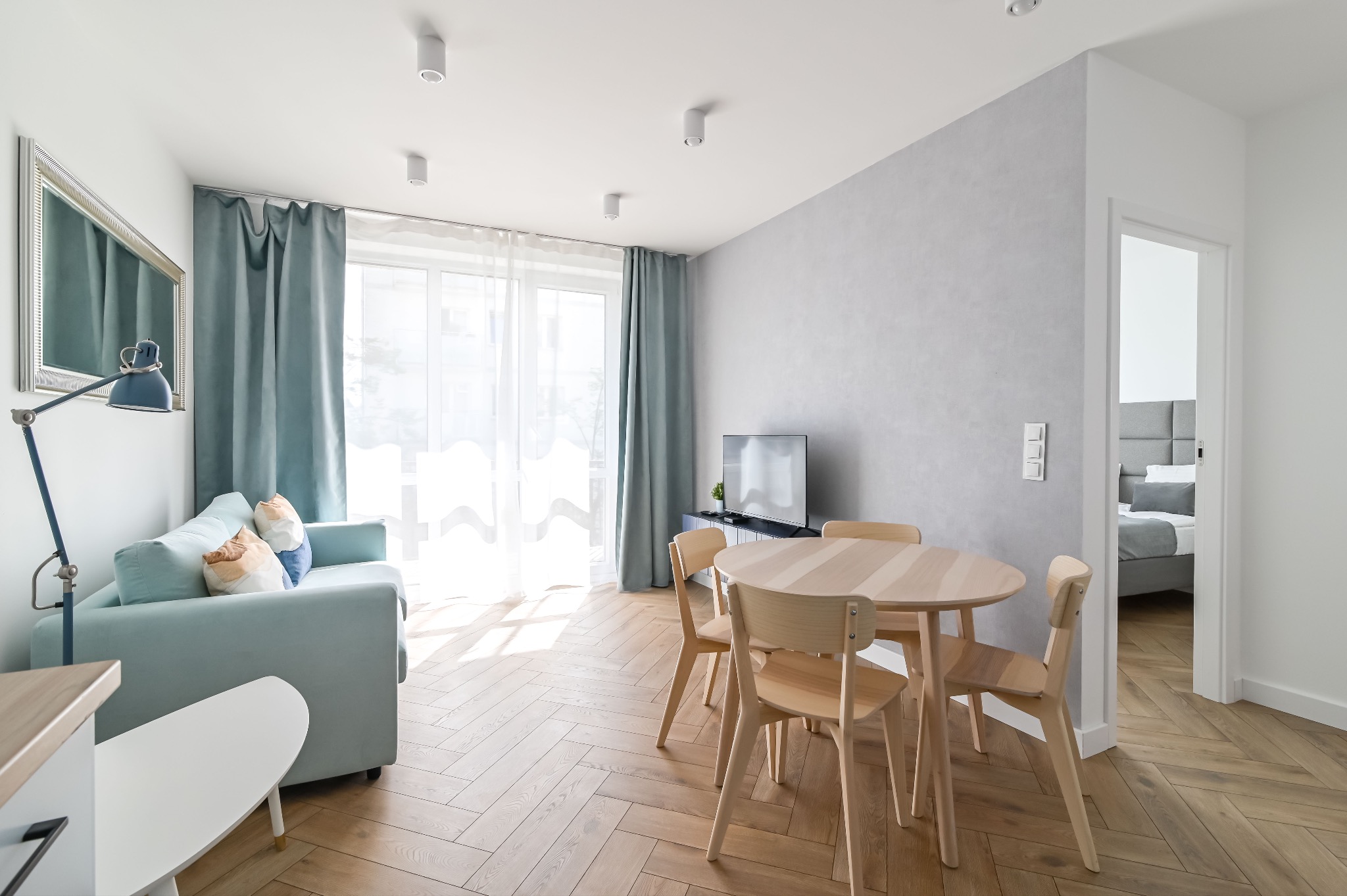 Jasne wnętrze: salon z parkietem w jodełkę, turkusową sofą i okrągłym stołem z krzesłami. Widok na sypialnię w tle. Minimalistyczny design.