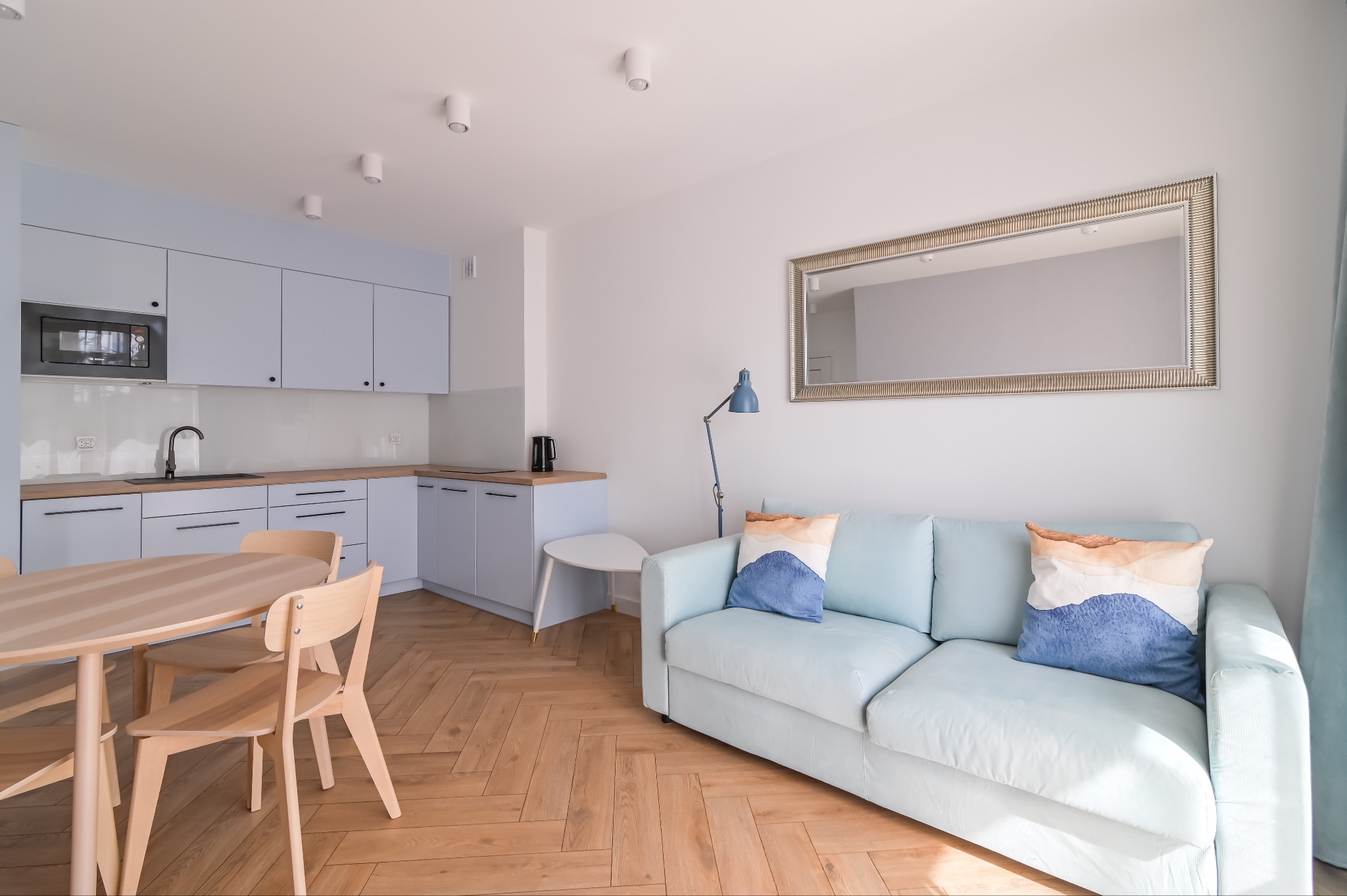 Jasne, przestronne wnętrze po remoncie: drewniana podłoga w jodełkę, błękitne meble kuchenne i wypoczynkowe, okrągły stół z krzesłami, lustro w ozdobnej ramie.