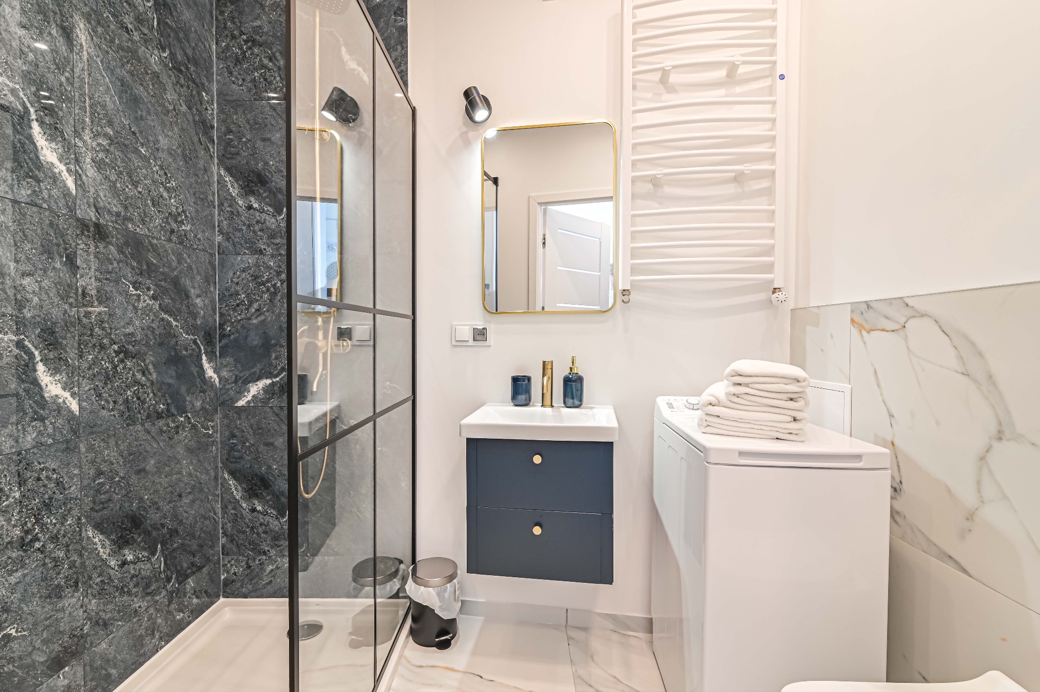 Elegancka łazienka po remoncie: kabina prysznicowa z czarnymi płytkami imitującymi marmur, szafka z umywalką w kolorze granatowym i biała pralka z ręcznikami.