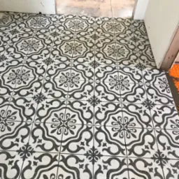 Ukończona podłoga z wzorzystych, czarno-białych płytek w łazience. Widoczne detale wzoru i precyzyjne ułożenie płytek. W tle fragmenty niedokończonych ścian.