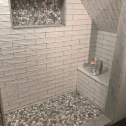 Wykończona kabina prysznicowa z białymi, podłużnymi płytkami i mozaiką z kamyków na podłodze i w niszy ściennej. Na półce leży zaprawa.