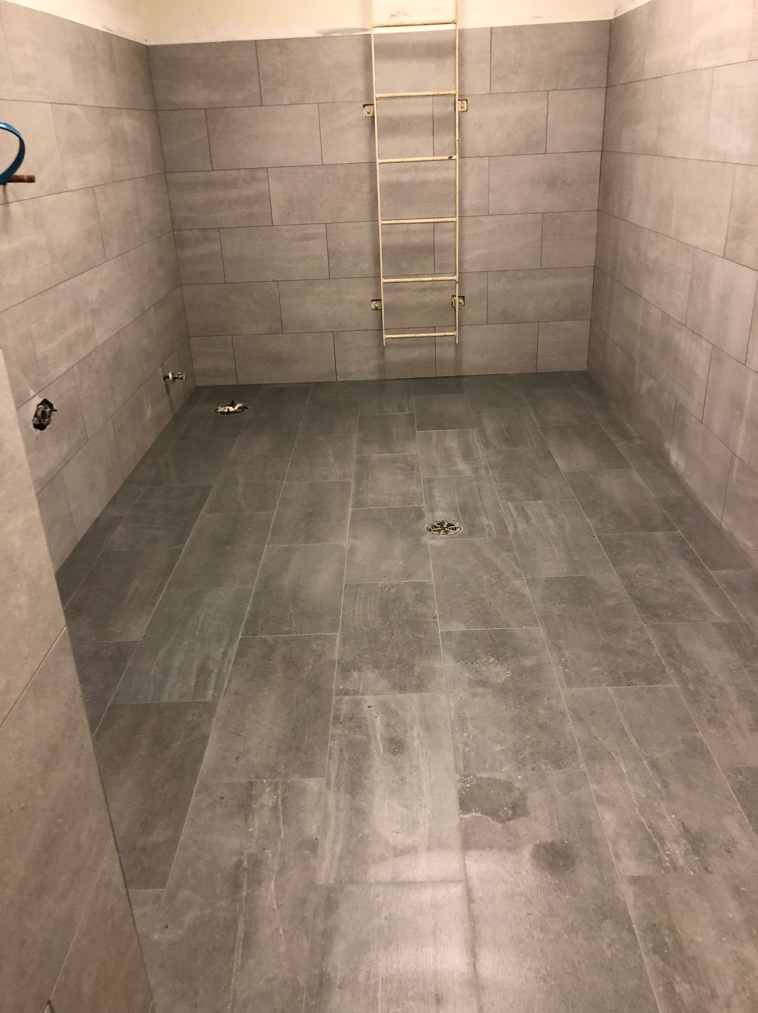 Wykończona kabina prysznicowa z szarymi płytkami imitującymi kamień, widoczny odpływ liniowy i drabinka na ścianie. Surowy, minimalistyczny styl.