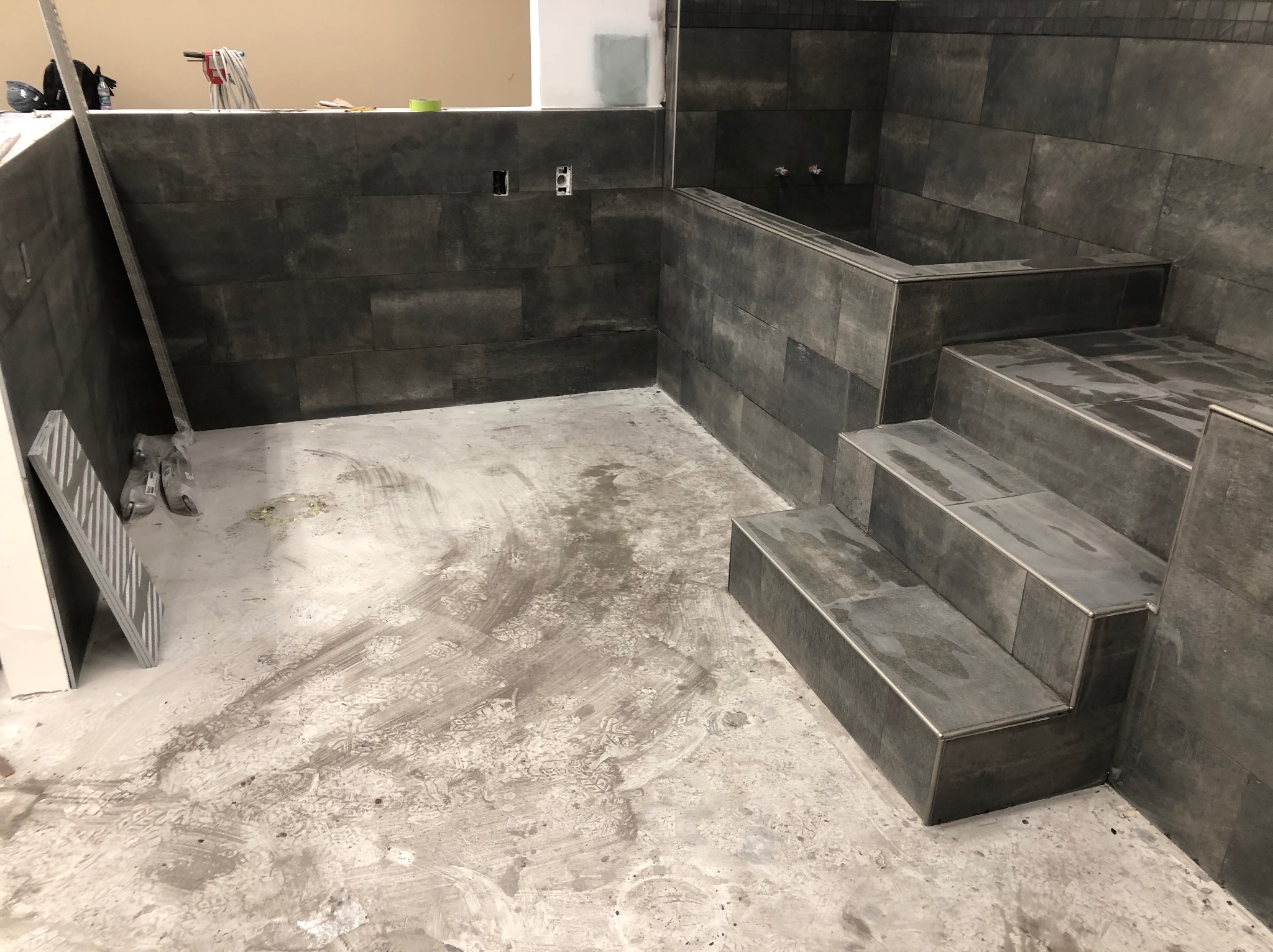 Wnętrze łazienki w trakcie wykańczania w Kętach. Widoczne ciemne płytki na ścianach i schodach wanny, surowy beton na podłodze. Etap prac glazurniczych.