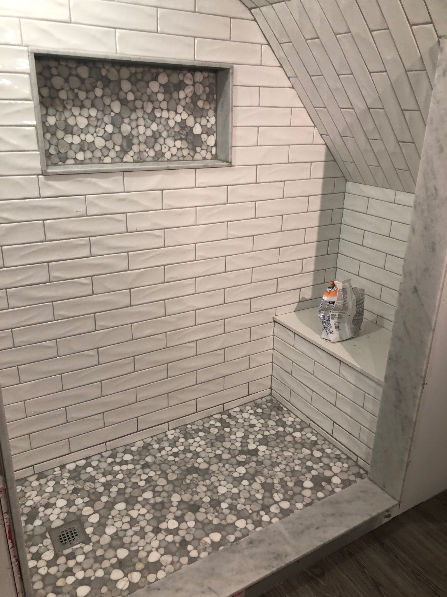 Wykończona kabina prysznicowa z białymi, podłużnymi płytkami i mozaiką z kamyków na podłodze i w niszy ściennej. Na półce leży zaprawa.