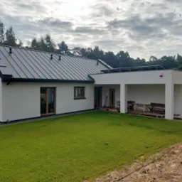 Nowoczesny dom z płaskim dachem i panelami słonecznymi, białą elewacją, ciemną blachą na dachu, widok z trawnika. Na zewnątrz drewniane meble ogrodowe.