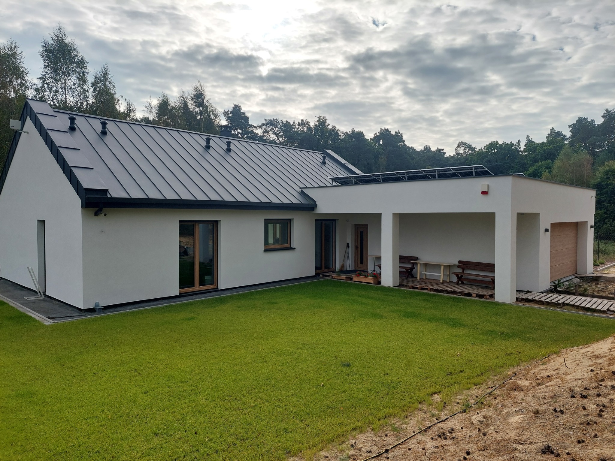 Nowoczesny dom z płaskim dachem i panelami słonecznymi, białą elewacją, ciemną blachą na dachu, widok z trawnika. Na zewnątrz drewniane meble ogrodowe.