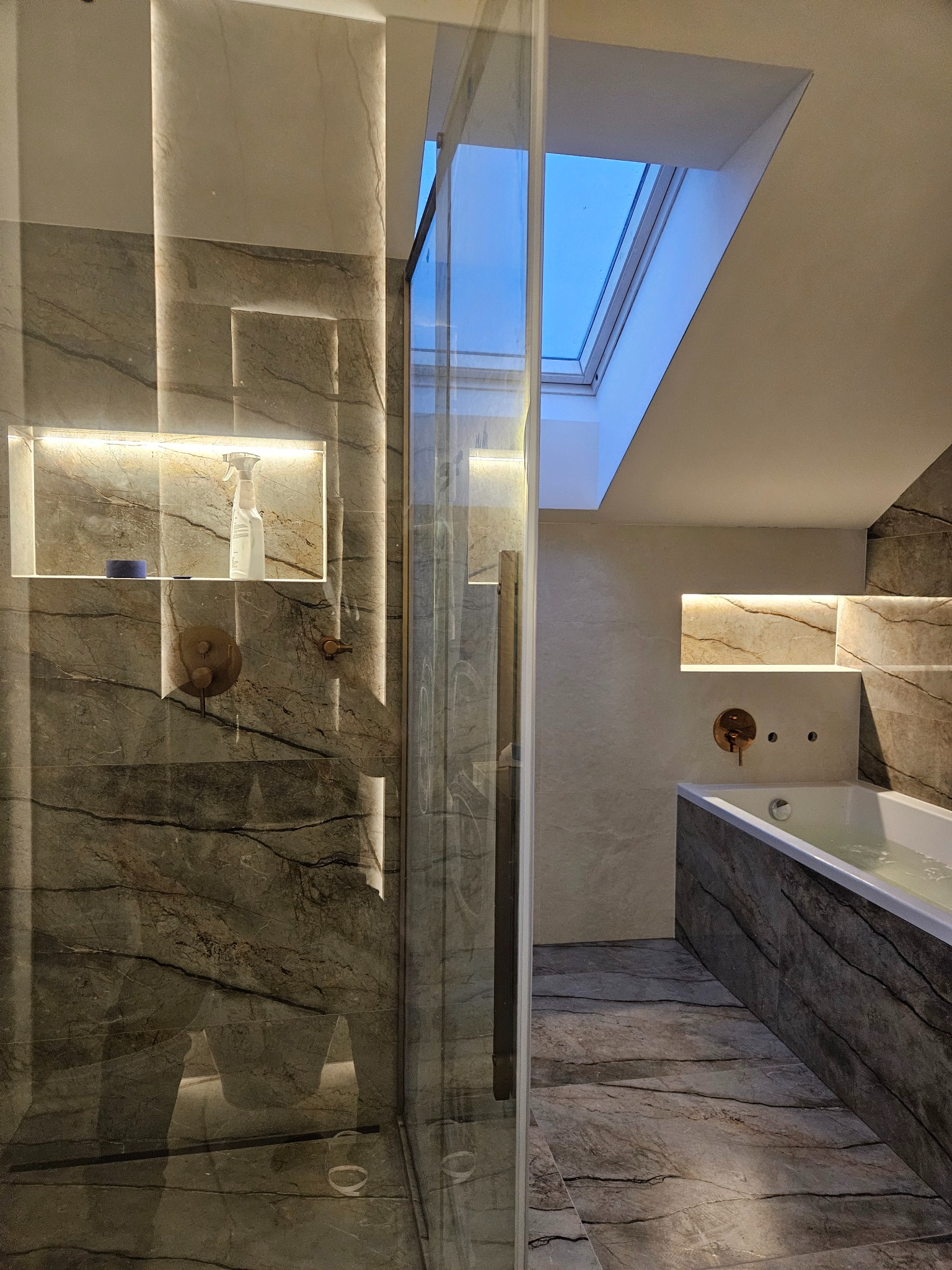 Ekskluzywna łazienka z marmurowymi płytkami, podświetlanymi niszami i szklanym prysznicem. Widok na okno dachowe.
