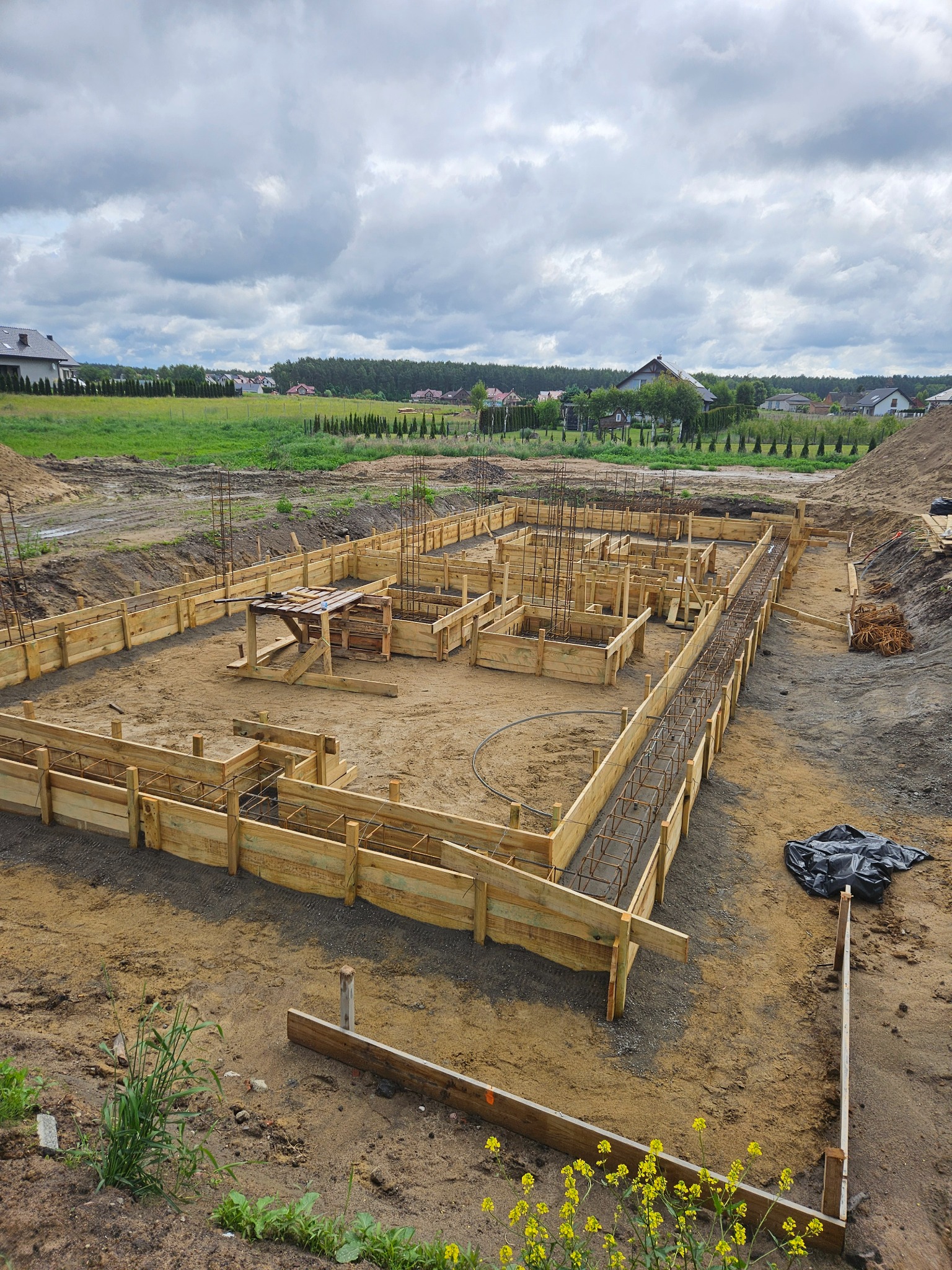 Szeroki kadr na przygotowany szalunek i zbrojenie fundamentów domu jednorodzinnego na działce budowlanej w słoneczny dzień. Widoczne elementy drewniane i stalowe.