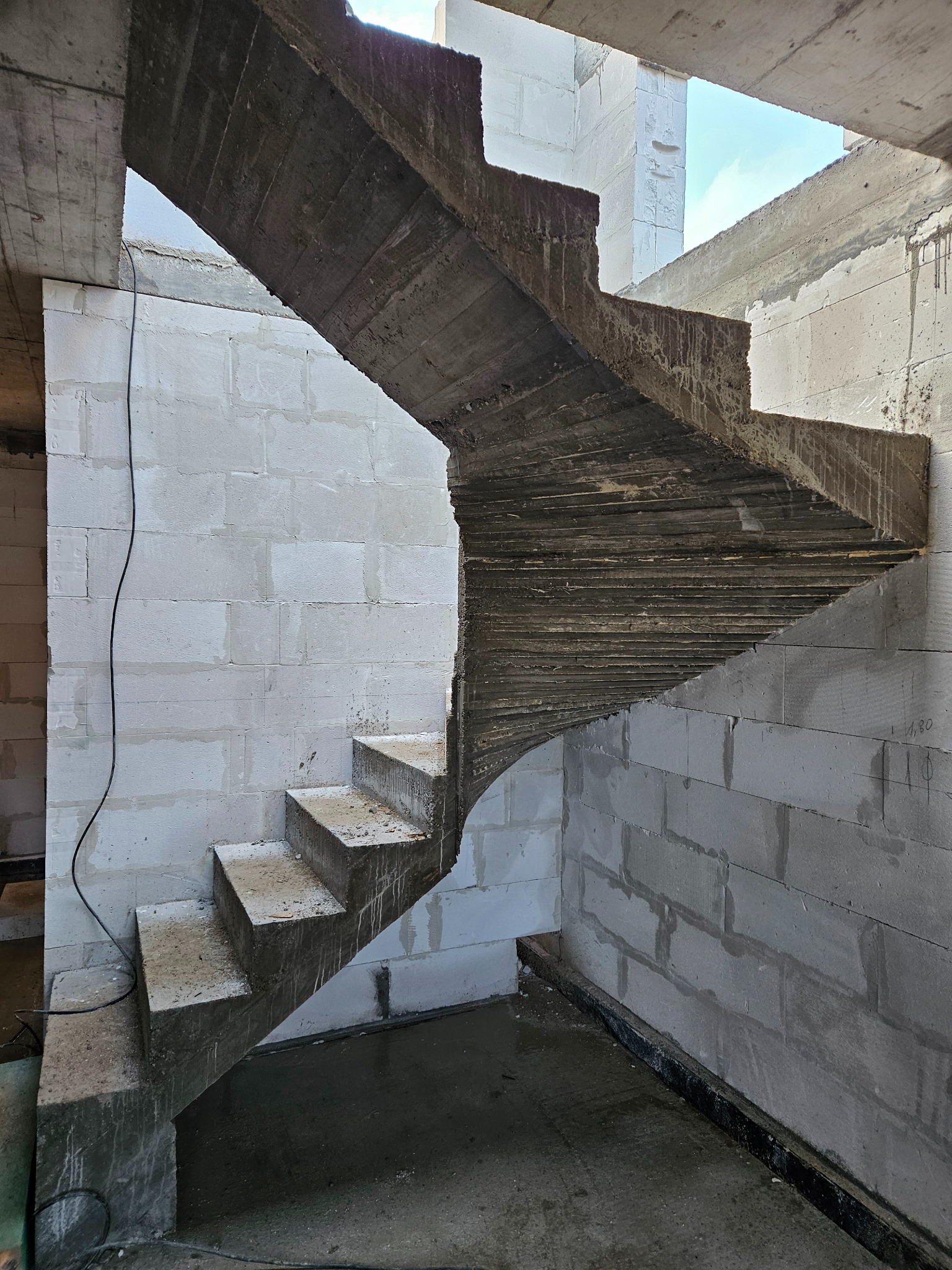 Surowe betonowe schody w trakcie budowy, widoczne białe ściany z bloczków, szare podłoże. W tle widoczne niebo przez otwór w konstrukcji.