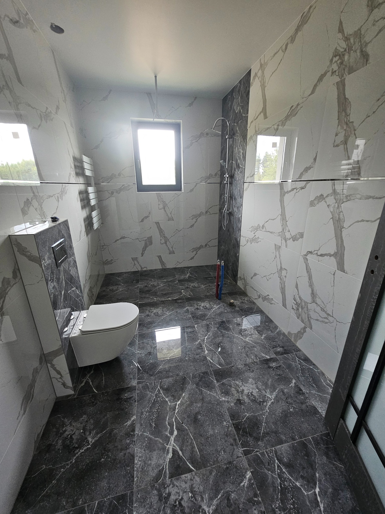 Wykończona łazienka z marmurowymi płytkami na ścianach i podłodze, widoczna toaleta, grzejnik drabinkowy i kabina prysznicowa z deszczownicą. Nowoczesny design.