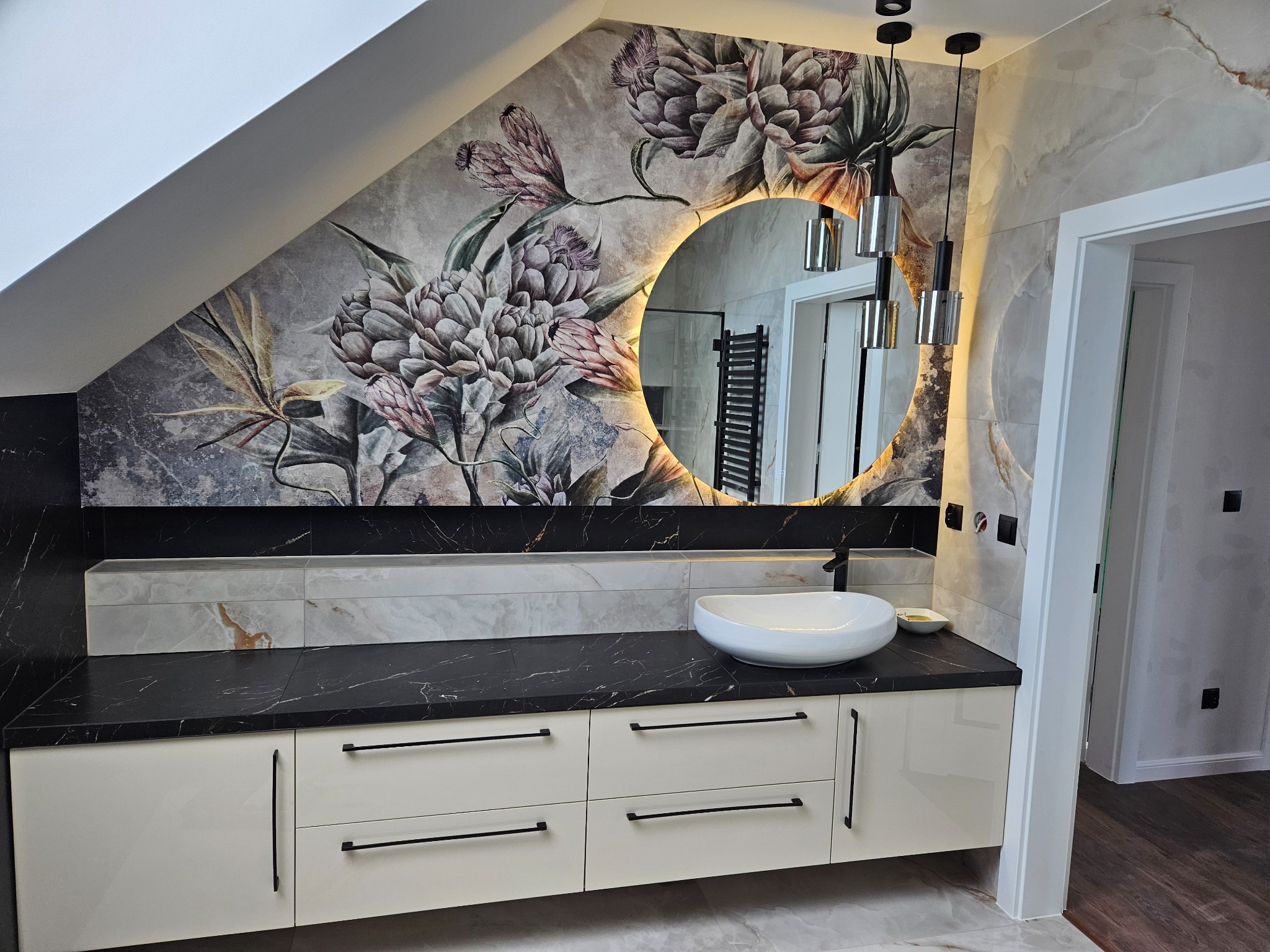 Elegancka łazienka na poddaszu z okrągłym lustrem podświetlanym LED, szafką z czarnym blatem i umywalką nablatową. Na ścianie tapeta w kwiaty.