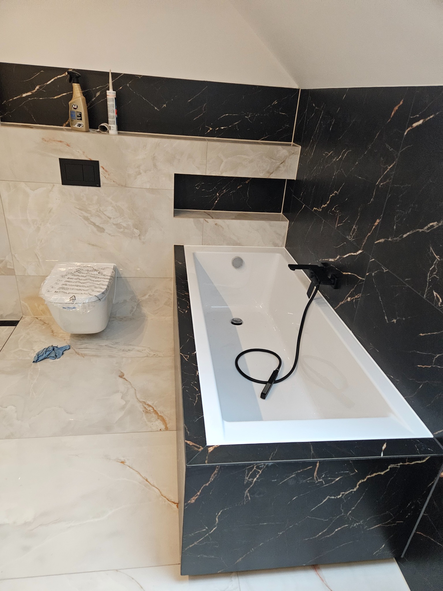 Nowoczesna łazienka z wanną obudowaną czarnym marmurem, białą ceramiką i beżowymi płytkami imitującymi marmur. Umywalka i toaleta w folii ochronnej.