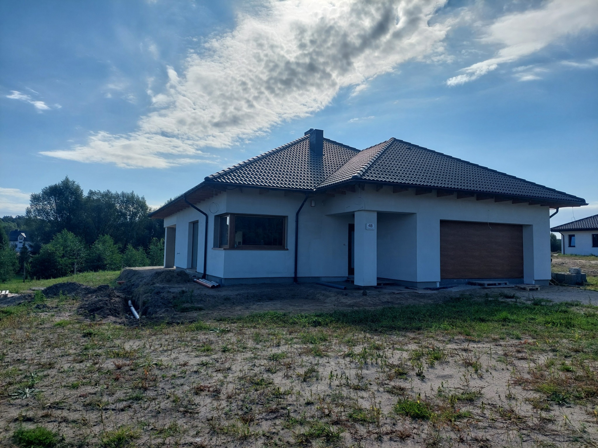 Niedawno wybudowany, parterowy dom z ciemną dachówką, jasną elewacją i drewnianą bramą garażową, otoczony piaszczystym terenem i zielenią. Widoczne elementy wykończeniowe.