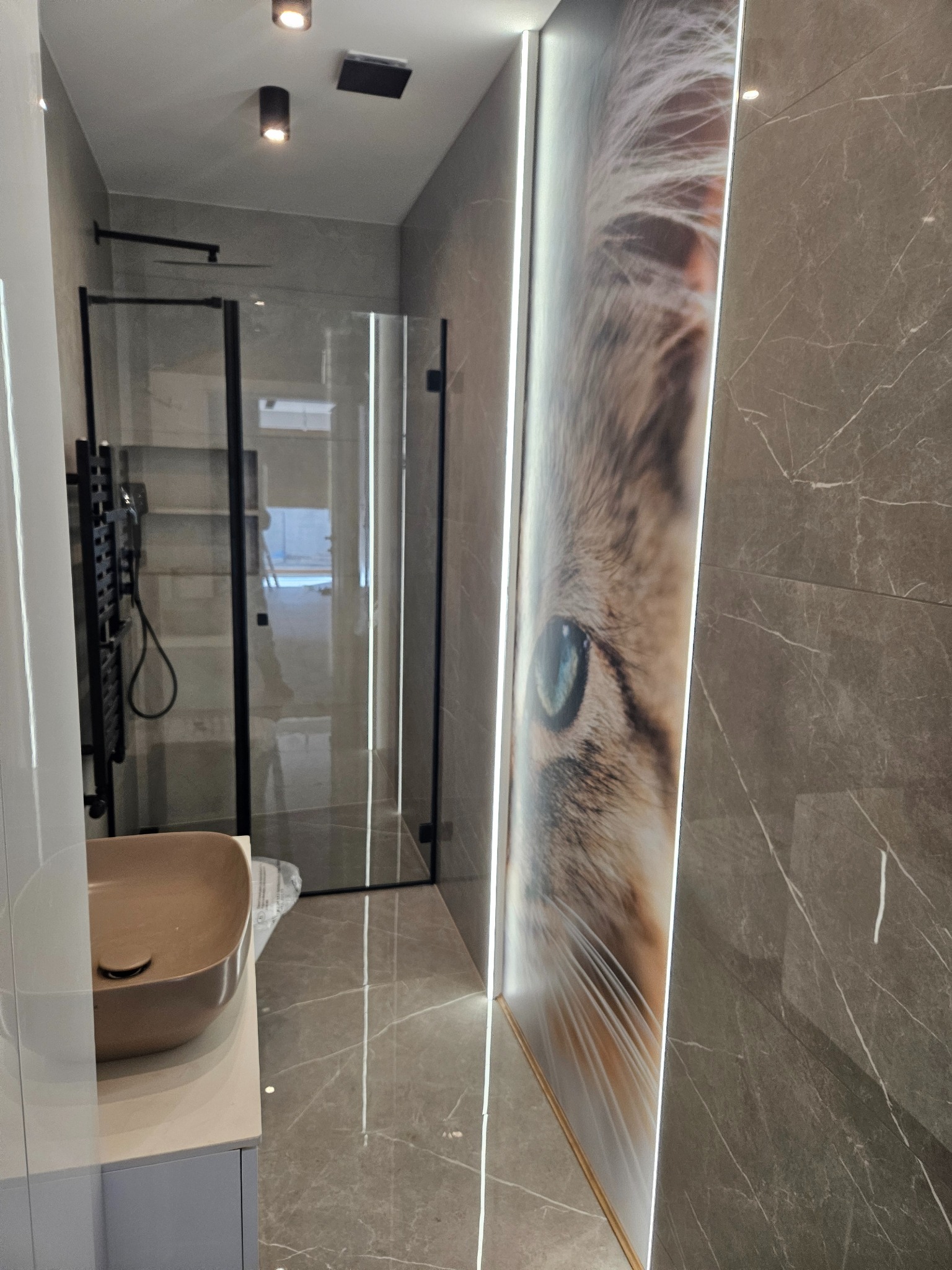 Nowoczesna łazienka z kabiną prysznicową typu walk-in, umywalką nablatową i podświetlaną grafiką z kocim okiem na ścianie z płytek imitujących marmur.