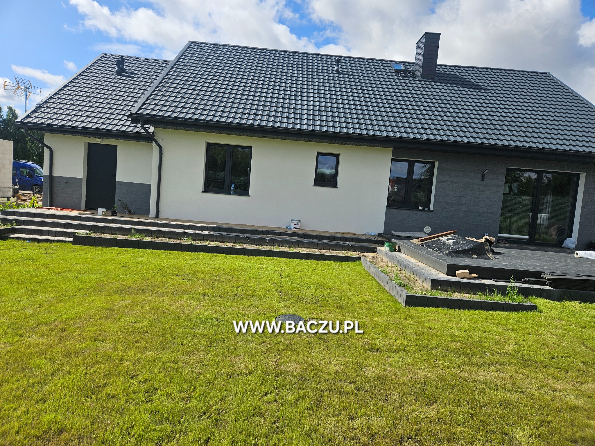 Nowoczesny dom jednorodzinny z ciemną dachówką i elementami szarej elewacji, widok z ogrodu, w tle błękitne niebo z chmurami. Widoczny fragment tarasu.