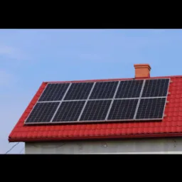 Panele słoneczne na czerwonym dachu domu jednorodzinnego, widoczne pod kątem. Kominy z cegły. Błękitne niebo w tle. Instalacja fotowoltaiczna.