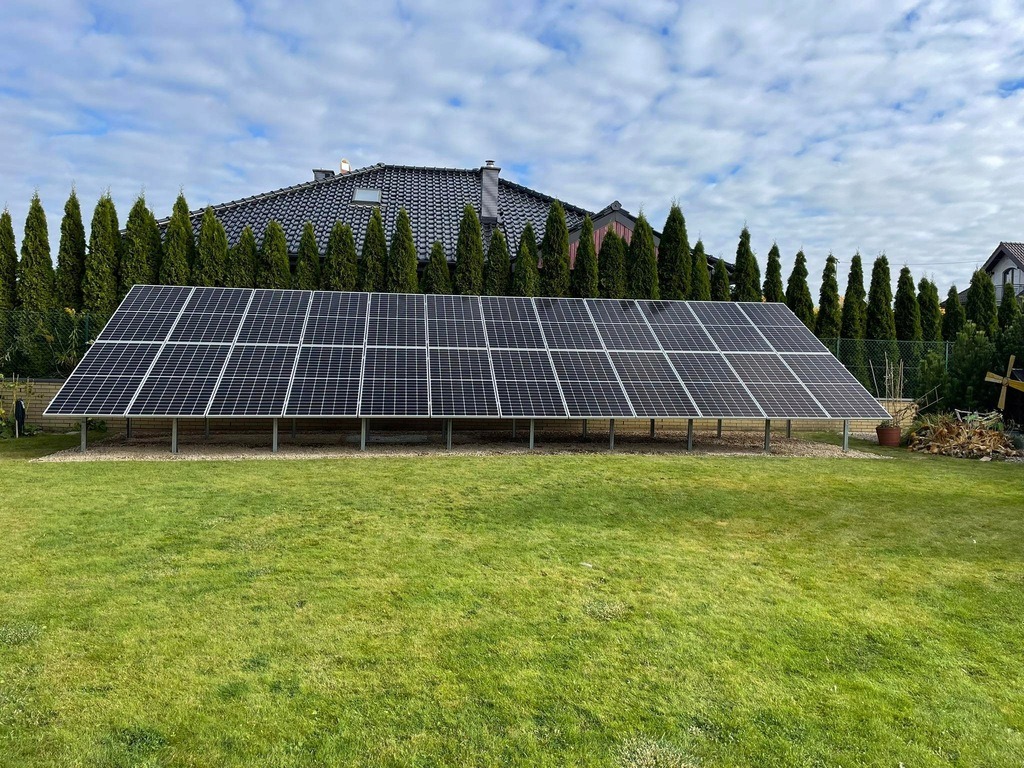 Panel fotowoltaiczny na trawniku, widok z przodu. W tle rząd tuj i dom. Instalacja na metalowej konstrukcji, panele w czarnych ramach. Słoneczny dzień.