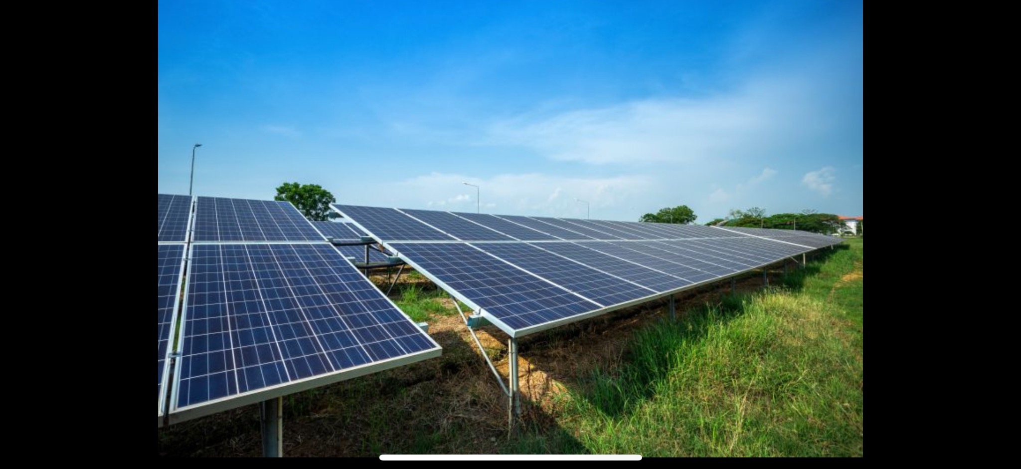Rząd paneli fotowoltaicznych na metalowej konstrukcji, na tle zielonej trawy i błękitnego nieba, oświetlonych jasnym światłem słonecznym.