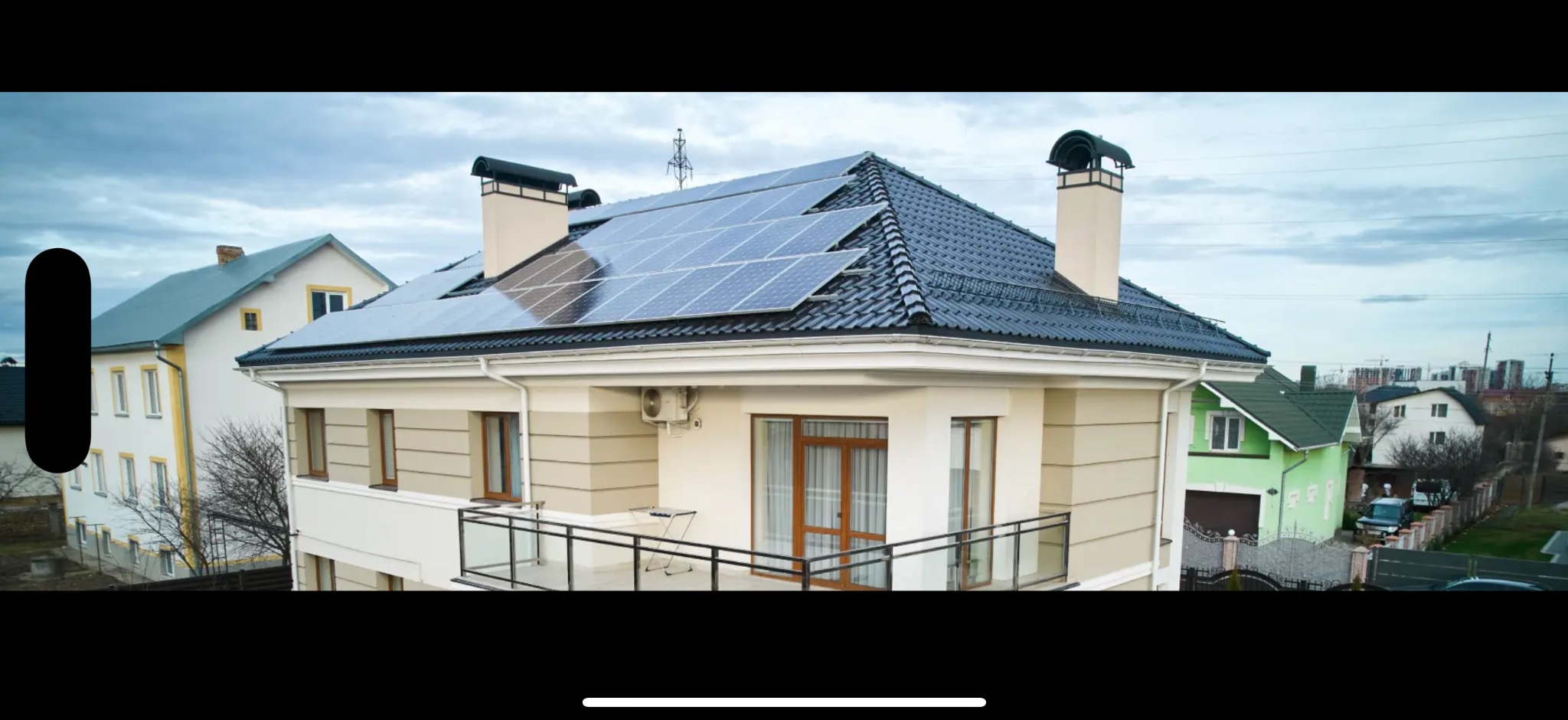 Dom jednorodzinny z panelami fotowoltaicznymi na czarnym dachu, widok z boku, na tle innych domów i pochmurnego nieba. Widoczny klimatyzator na ścianie budynku.