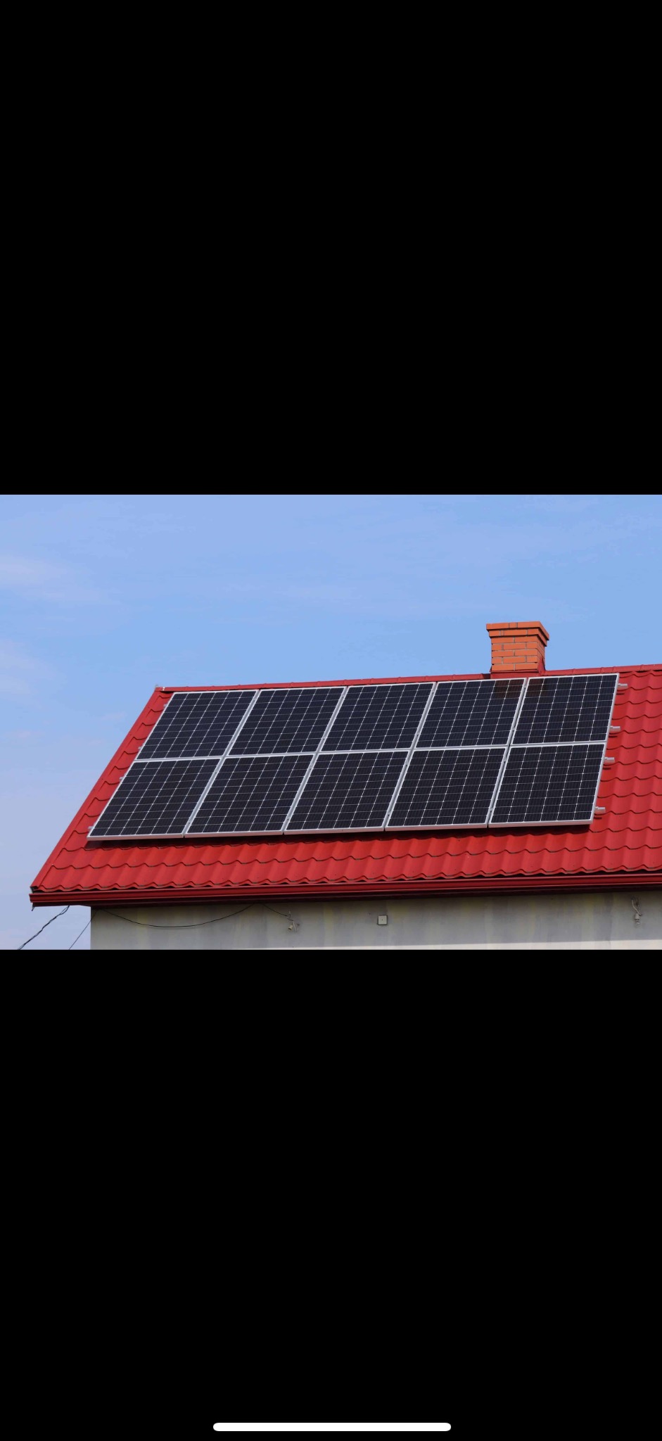Panele słoneczne na czerwonym dachu domu jednorodzinnego, widoczne pod kątem. Kominy z cegły. Błękitne niebo w tle. Instalacja fotowoltaiczna.