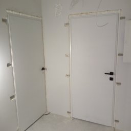 Przemysław Waluś - Dwa białe, zamontowane drzwi wewnętrzne z czarnymi klamkami w surowym wnętrzu. Widoczne elementy montażowe i przewody elektryczne.