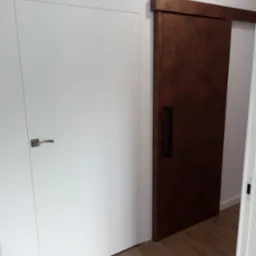 Dwa rodzaje drzwi: białe drzwi z klamką i ciemnobrązowe drzwi przesuwne z długim uchwytem, na tle drewnianej podłogi i białych ścian.