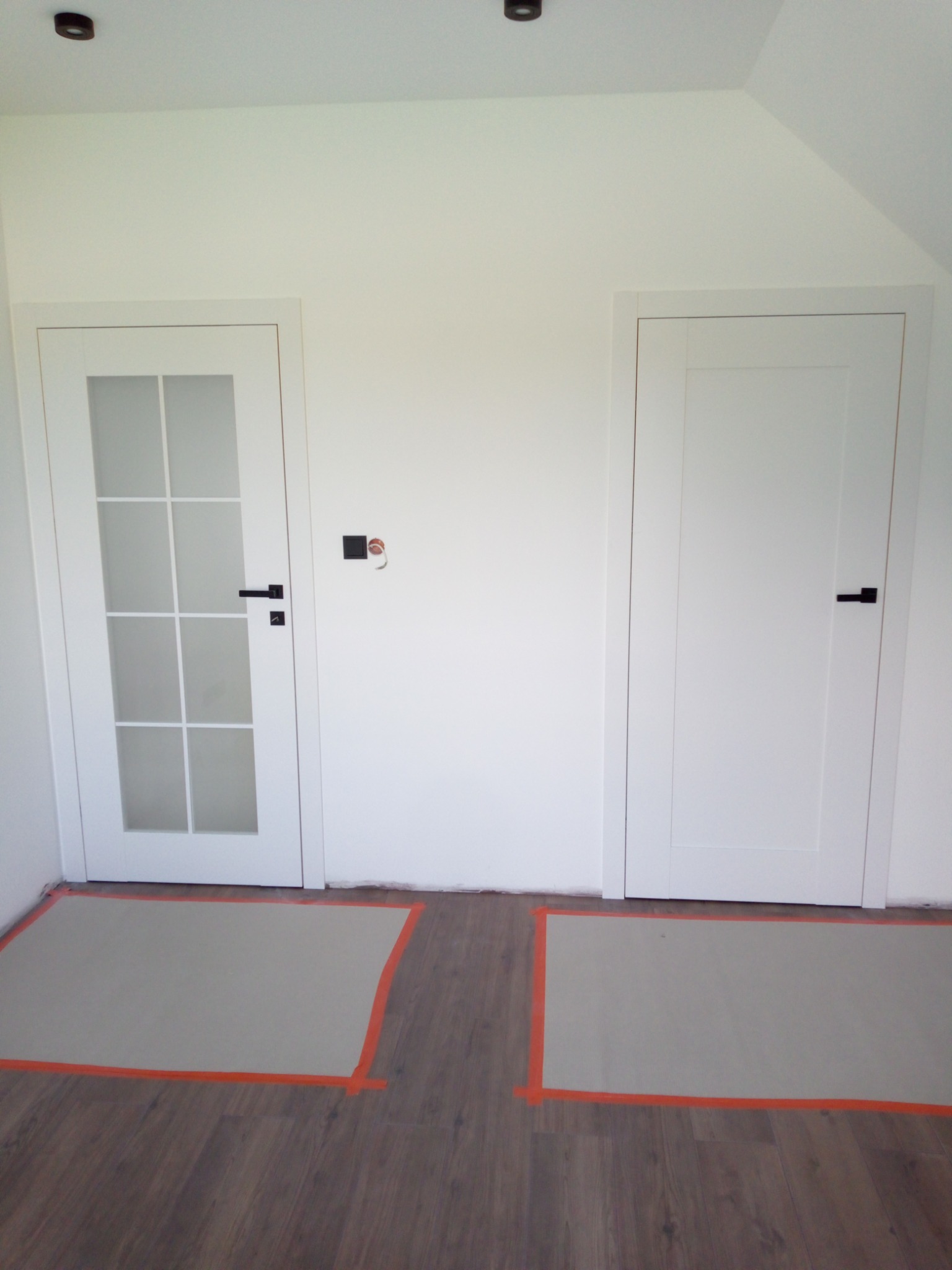 Dwie białe, nowoczesne drzwi z czarnymi klamkami w jasnym wnętrzu. Jedne drzwi przeszklone, drugie pełne. Podłoga zabezpieczona kartonem i taśmą malarską.