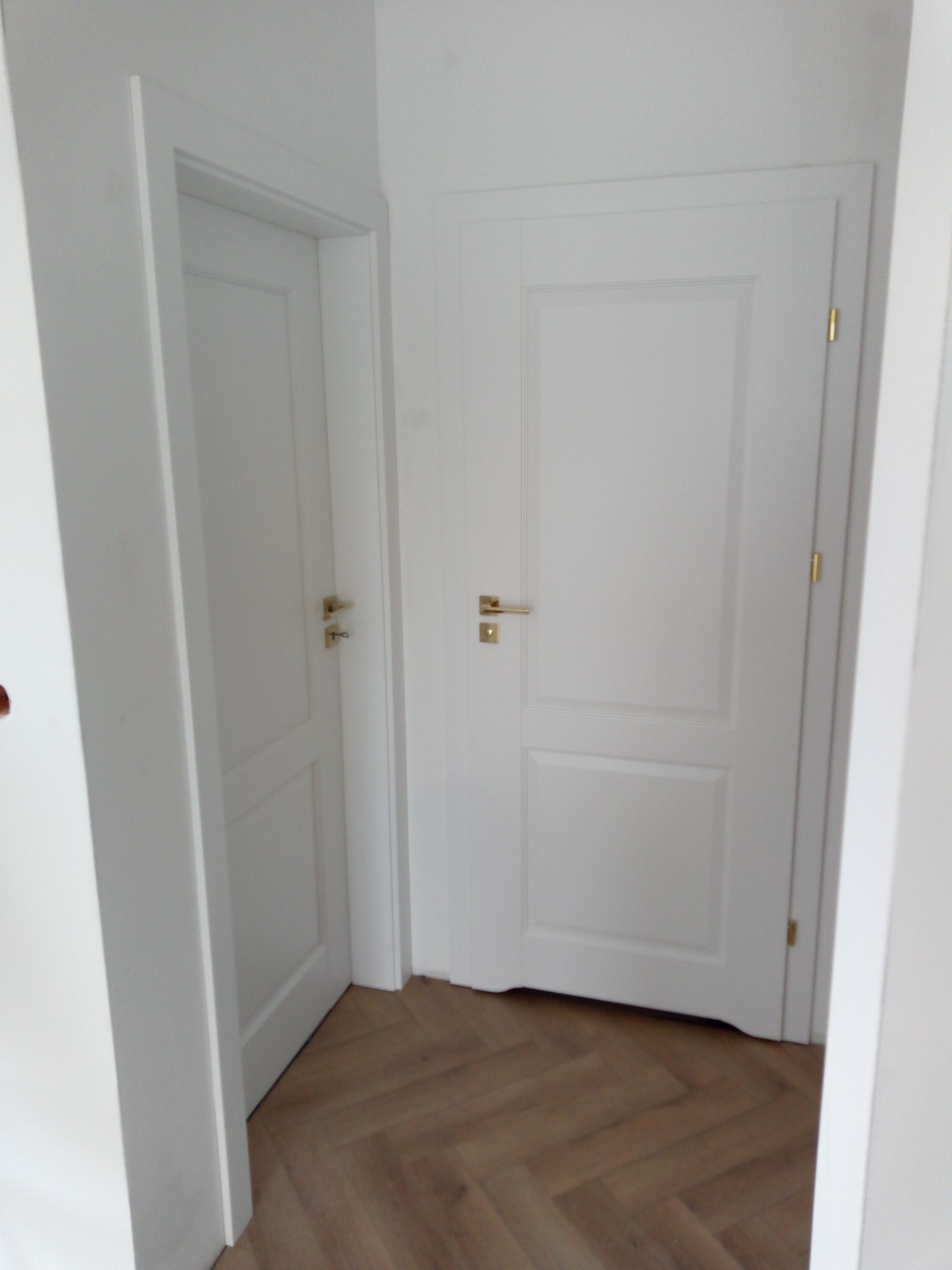 Dwa białe, klasyczne drzwi wewnętrzne z złotymi klamkami, zamontowane w białej ścianie, widoczne na tle parkietu ułożonego w jodełkę.