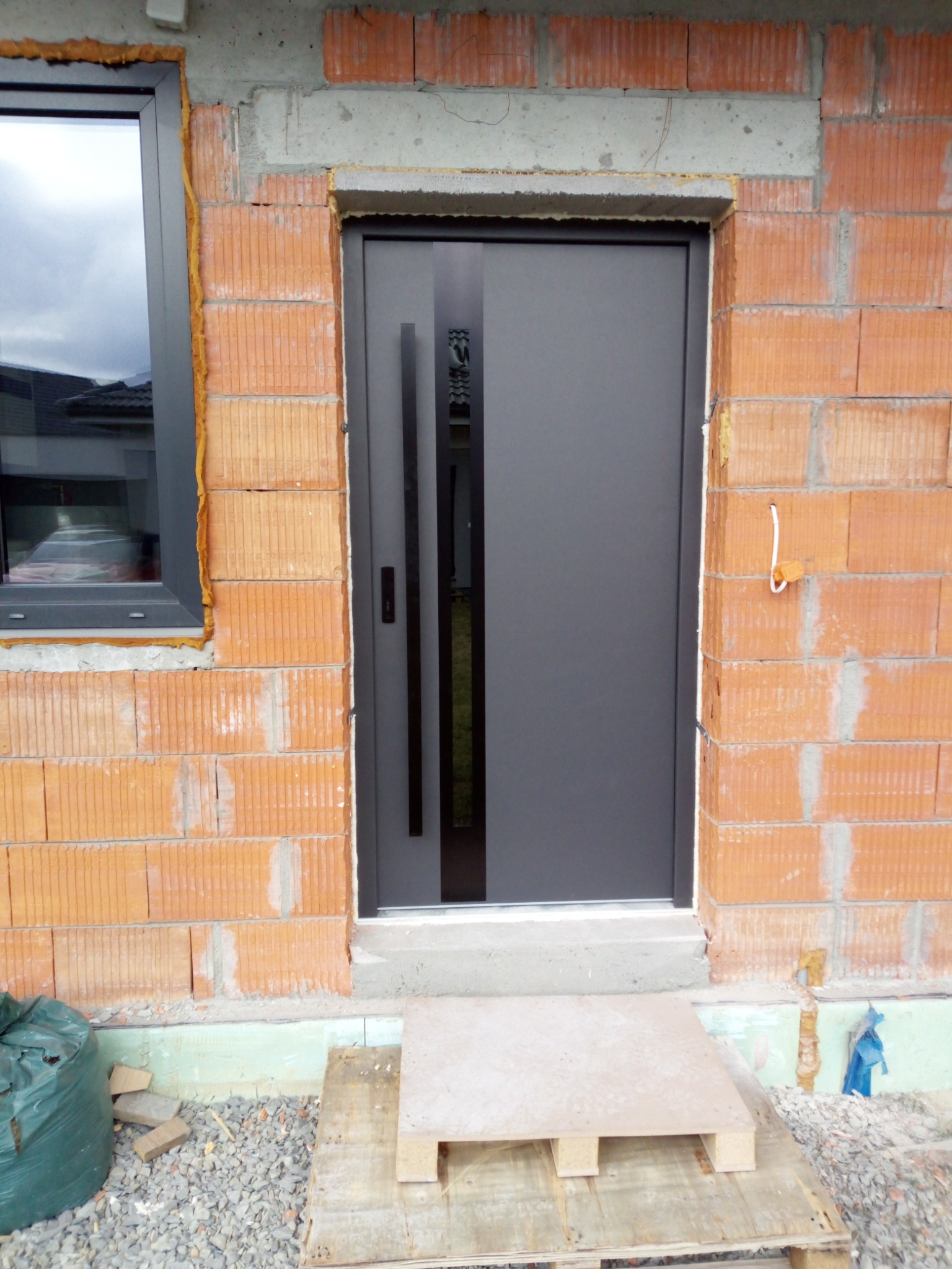 Nowoczesne, grafitowe drzwi wejściowe z długim uchwytem, zamontowane w surowym murze z cegły ceramicznej. Widoczne okno i fragment ocieplenia.