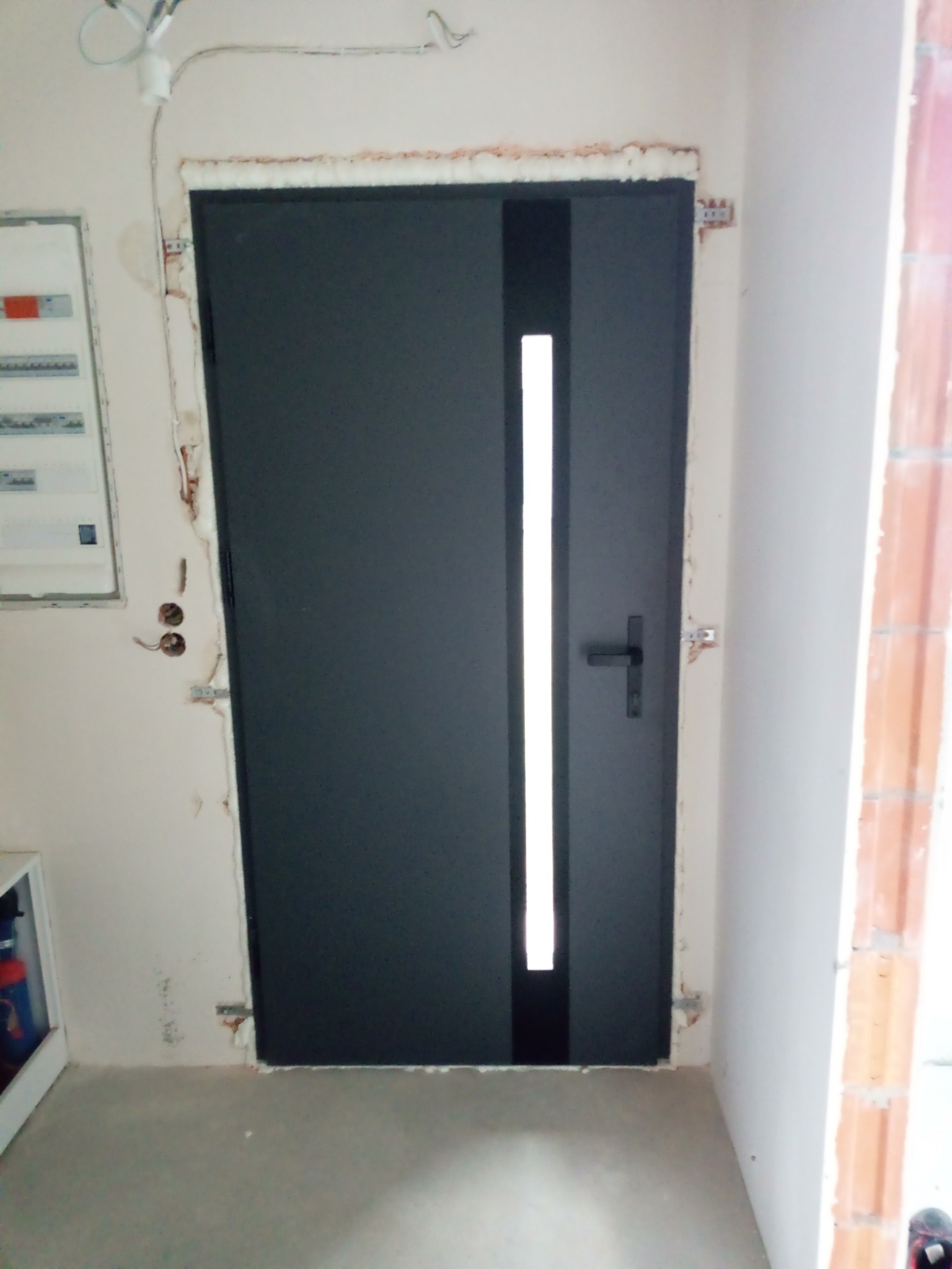 Ciemne drzwi z pionowym przeszkleniem, zamontowane w surowym otworze, widoczne ślady po montażu na ścianie. Nowe drzwi w trakcie wykańczania.