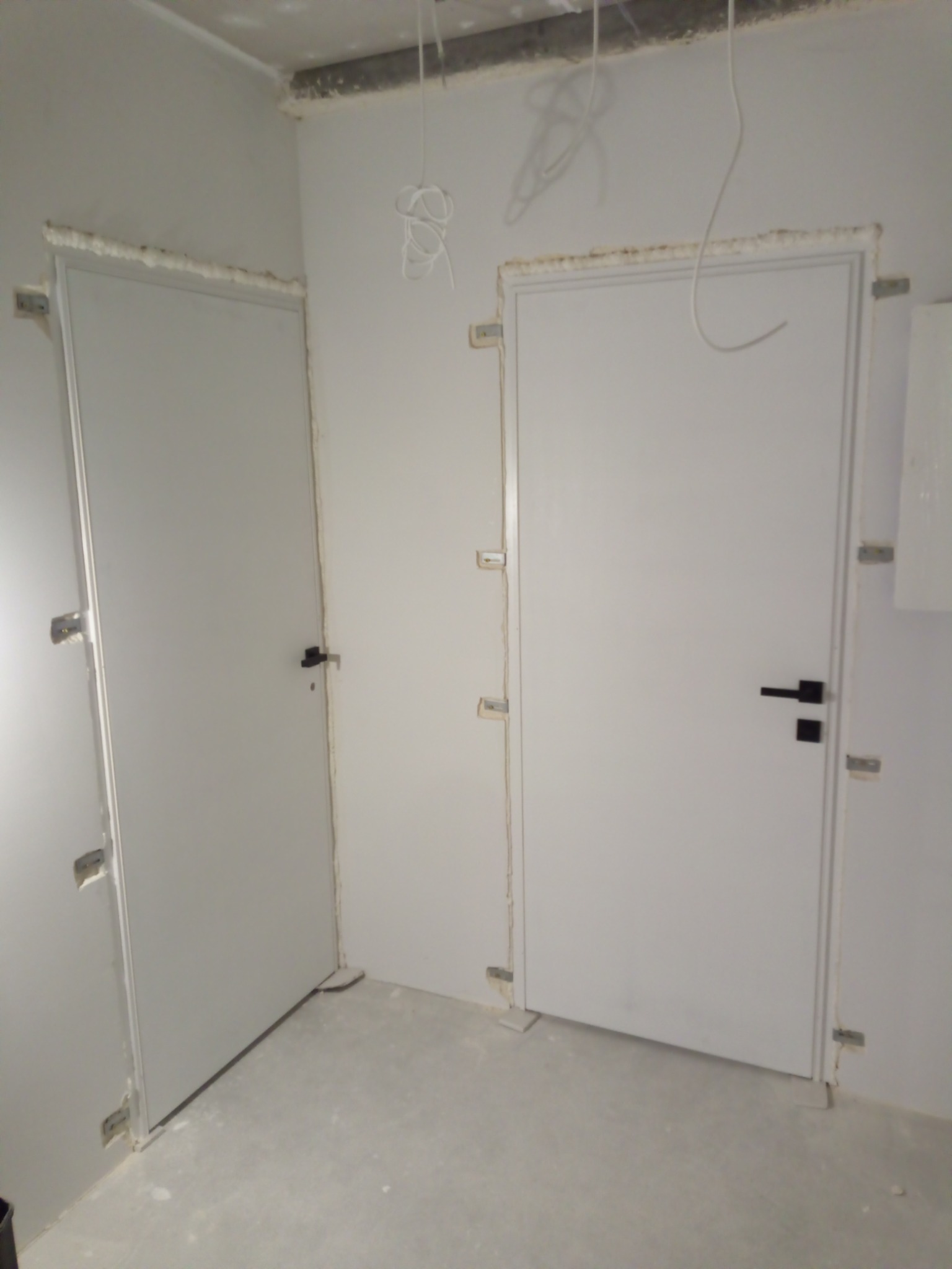 Dwa białe, zamontowane drzwi wewnętrzne z czarnymi klamkami w surowym wnętrzu. Widoczne elementy montażowe i przewody elektryczne.