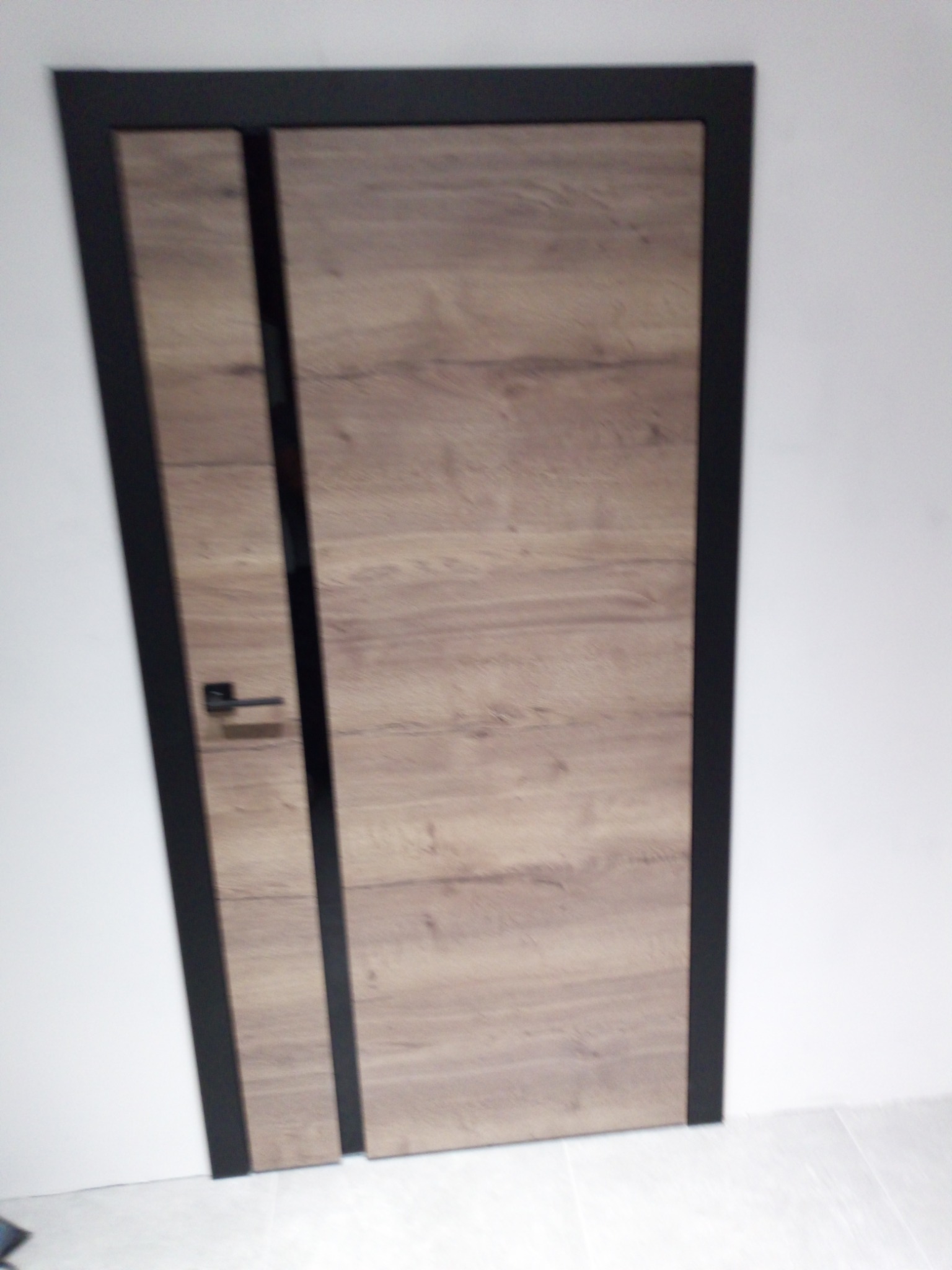 Drewnopodobne drzwi z czarną ramą i klamką, zamontowane w białej ścianie. Widoczne wykończenie i detale.