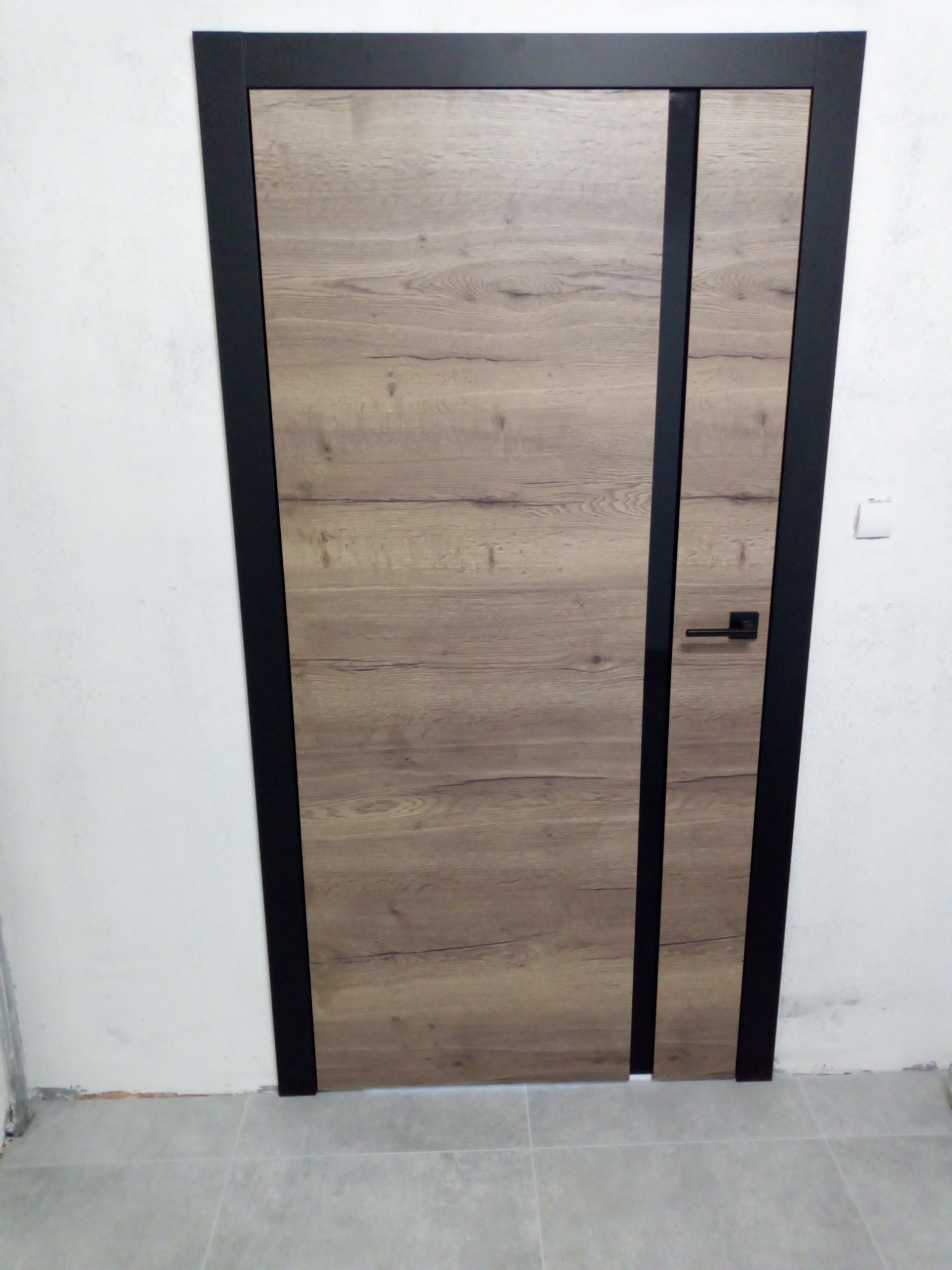 Zamknięte drzwi wewnętrzne z okleiną drewnopodobną i czarną ramą, widoczne na tle białej ściany i szarej podłogi.