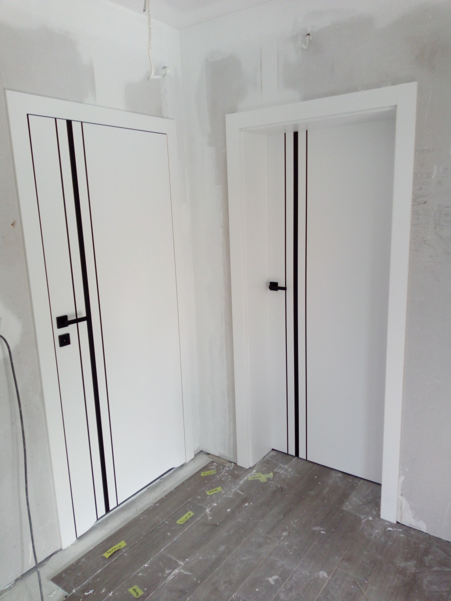 Dwa białe, nowoczesne drzwi z czarnymi paskami i klamkami, zamontowane w surowym wnętrzu z szarą podłogą, w trakcie prac wykończeniowych.