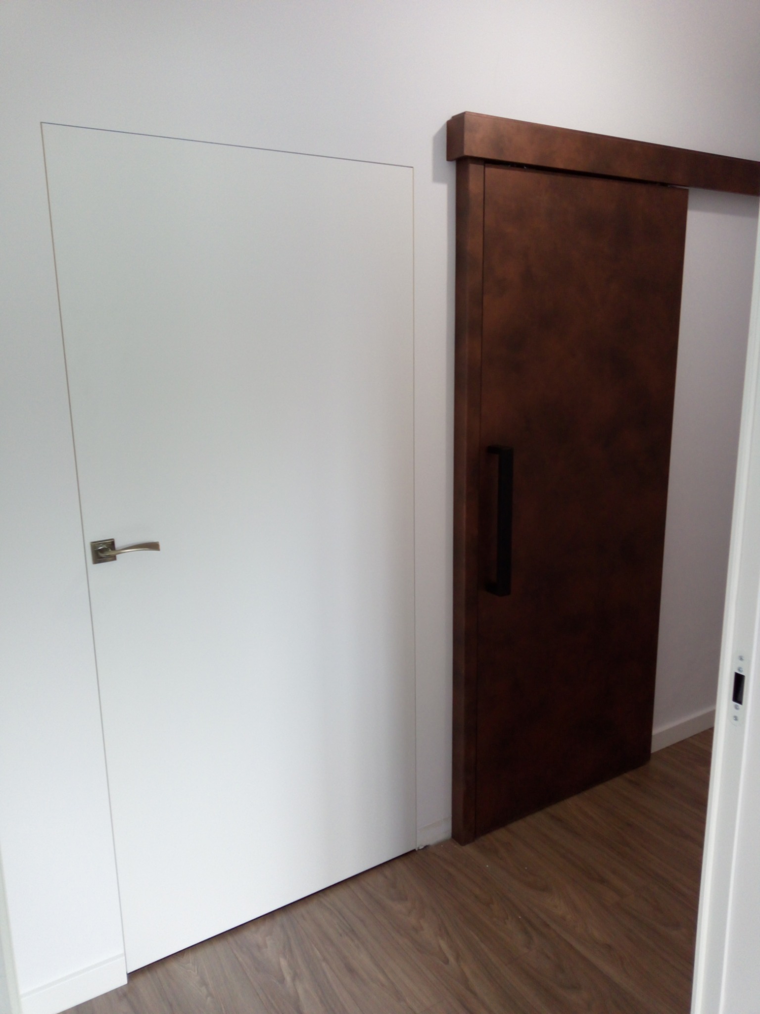 Dwa rodzaje drzwi: białe drzwi z klamką i ciemnobrązowe drzwi przesuwne z długim uchwytem, na tle drewnianej podłogi i białych ścian.