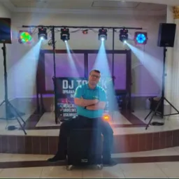 DJ Tomek siedzi na głośniku, otoczony sprzętem nagłośnieniowym i oświetleniowym na sali weselnej. Widoczne reflektory i kolumny głośnikowe.