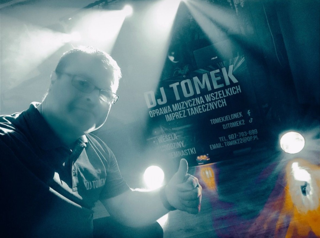 DJ Tomek prezentuje się na tle baneru reklamowego, dając kciuk w górę. Scena oświetlona reflektorami, z widocznymi danymi kontaktowymi.