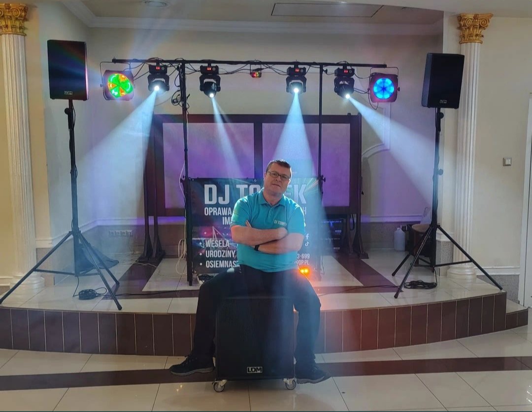 DJ Tomek siedzi na głośniku, otoczony sprzętem nagłośnieniowym i oświetleniowym na sali weselnej. Widoczne reflektory i kolumny głośnikowe.
