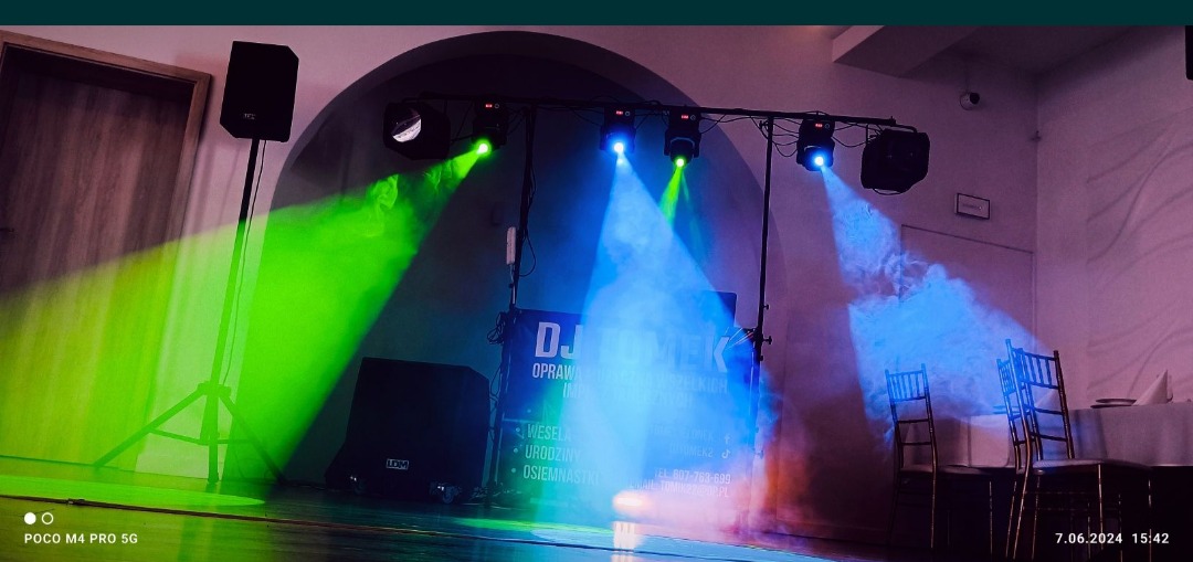 Scena z nagłośnieniem i oświetleniem DJ-a, zielone i niebieskie światła, sprzęt LDM, gotowa do imprezy w sali z nakrytymi stołami.