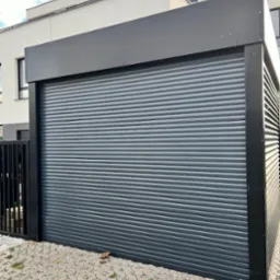 Nowoczesna, szara brama garażowa segmentowa z czarną ramą, zamontowana przy jasnym budynku. Kostka brukowa na podłożu.