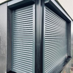 WIK. Rolety Zewnętrzne Szczecin. Bramy Rolowane. Bramy garażowe. Kraty rolowane - Szare rolety zewnętrzne na dwóch oknach narożnych, z widocznym systemem montażowym i fragmentem elewacji budynku. Nowoczesny design, minimalistyczny styl.