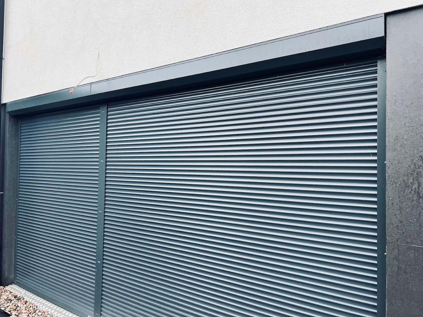 Zamknięta, szara roleta zewnętrzna z widocznymi lamelami, zamontowana na budynku z jasną elewacją. Widoczne mocowania i fragment elewacji.
