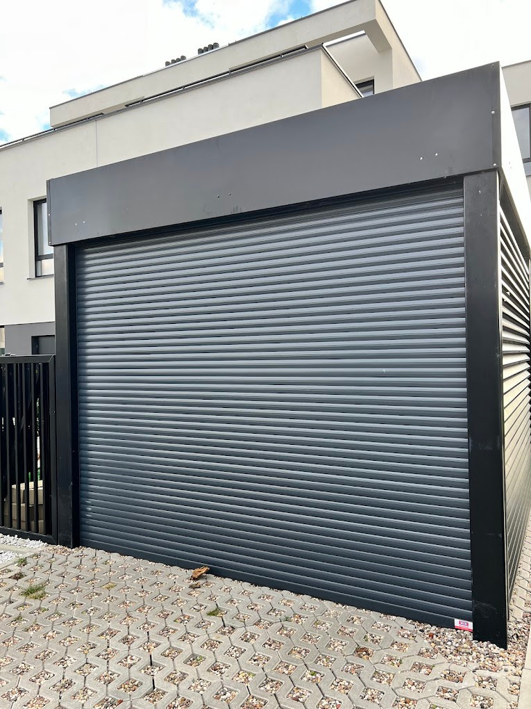 Nowoczesna, szara brama garażowa segmentowa z czarną ramą, zamontowana przy jasnym budynku. Kostka brukowa na podłożu.