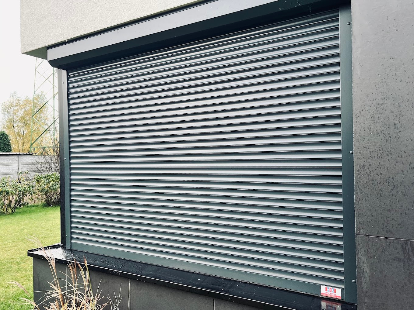 Zewnętrzna roleta okienna w kolorze grafitowym, zamontowana na tle nowoczesnej elewacji i zielonego trawnika. Widoczne detale mocowania i poziome lamele.