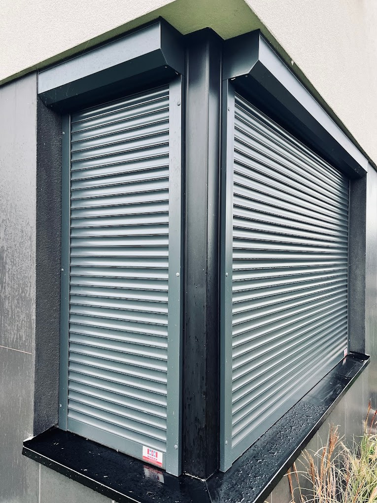 Szare rolety zewnętrzne na dwóch oknach narożnych, z widocznym systemem montażowym i fragmentem elewacji budynku. Nowoczesny design, minimalistyczny styl.