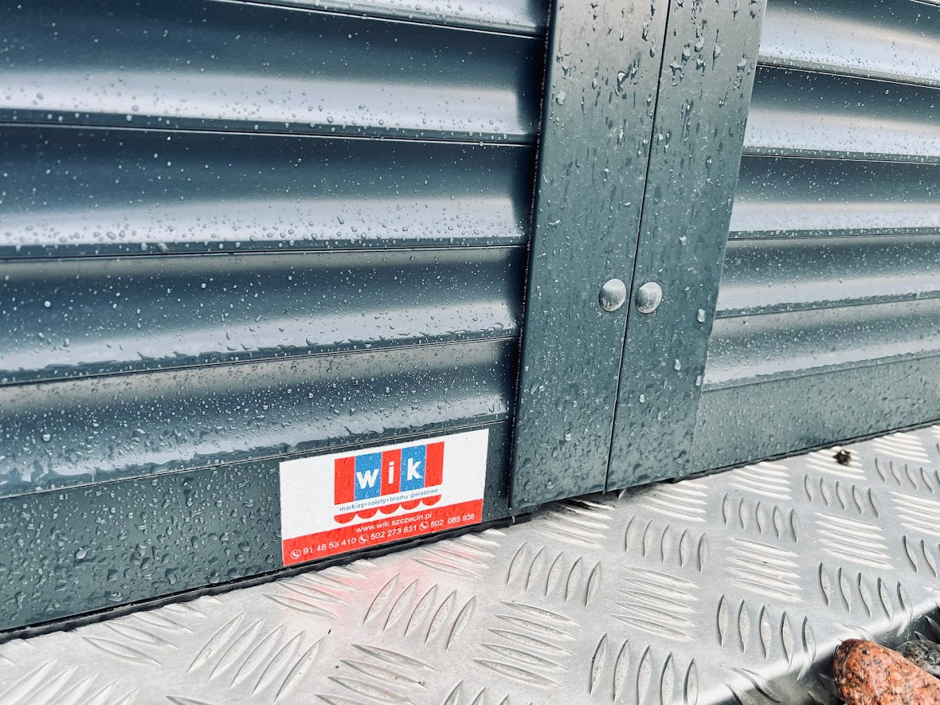 Szara, metalowa roleta zewnętrzna z widocznymi kroplami deszczu, zamontowana na metalowej powierzchni. Widoczne logo firmy z danymi kontaktowymi.
