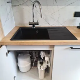 Czarny zlew granitowy z chromowaną baterią i widocznym systemem filtracji wody pod spodem, umieszczonym w szafce z białymi frontami.