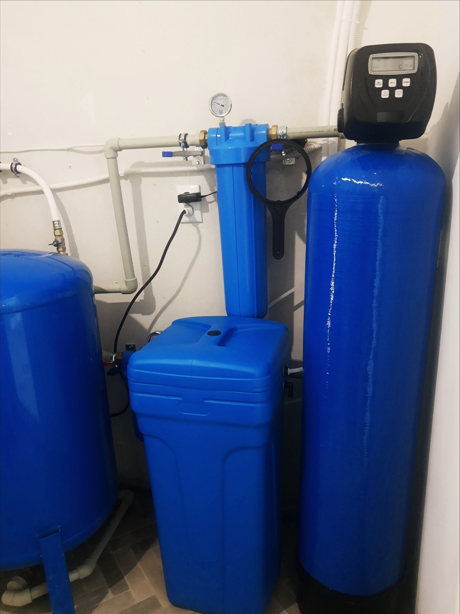 Niebieskie filtry do wody w pomieszczeniu, z manometrem i czarnym panelem sterowania, podłączone rurami do instalacji. Widoczny klucz do filtra.